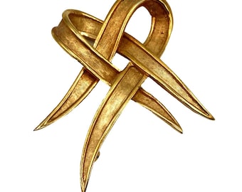 Pode incluir: Um broche de metal dourado em forma de nó ou fita estilizada. O broche tem uma superfície texturizada e um design pontiagudo.
