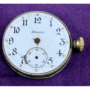 May include: Antique Illinois pocket watch face with a white dial, black numerals, and a subdial. The watch face has a gold-colored bezel and a winding stem. The dial is cracked, and the word "Illinois" is printed on the face.