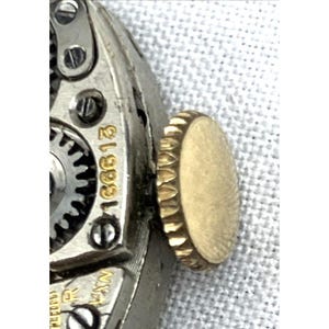 Op de afbeelding: Close-up van een vintage horlogemechanisme. Het horloge heeft zilverkleurige metalen componenten, waaronder tandwielen en schroeven. Een goudkleurige kroon is zichtbaar aan de rechterkant. Het nummer "168613" is op het mechanisme gegraveerd.