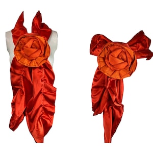 May include: A vibrant orange satin accessory featuring a large, layered rose in the center. The accessory has flowing, draped fabric and is displayed on a mannequin. The rose has a darker orange center.