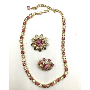 May include: A vintage jewelry set featuring a gold-toned necklace and two brooches. The necklace and brooches are adorned with pink and clear rhinestones. One brooch is a flower design with faux pearls, and the other is a stylized design with pink stones.