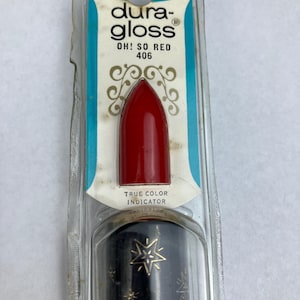 May include: A Dura-Gloss lipstick in the shade Oh! So Red 406, encased in original clear plastic packaging. The product features a red color indicator at the top and a black tube decorated with gold star patterns, with the brand name printed clearly.