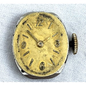 Op de afbeelding: Een vintage uurwerk met een goudkleurige wijzerplaat en wijzers, met zwarte cijfers op de posities 1, 3, 6, 9 en 12 uur. Het horloge heeft een zilverkleurige metalen behuizing en een zwarte opwindkroon aan de zijkant. De wijzerplaat vertoont tekenen van slijtage.