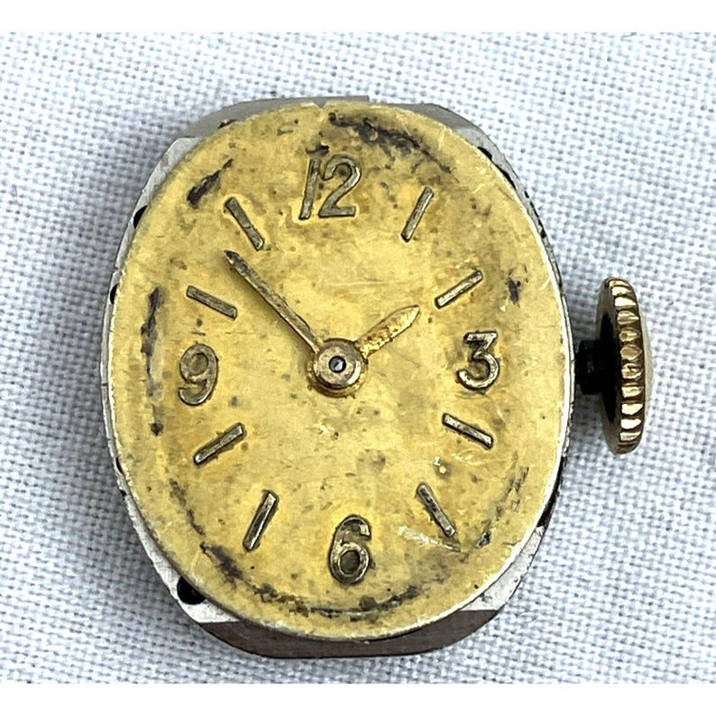 Op de afbeelding: Een vintage uurwerk met een goudkleurige wijzerplaat en wijzers, met zwarte cijfers op de posities 1, 3, 6, 9 en 12 uur. Het horloge heeft een zilverkleurige metalen behuizing en een zwarte opwindkroon aan de zijkant. De wijzerplaat vertoont tekenen van slijtage.