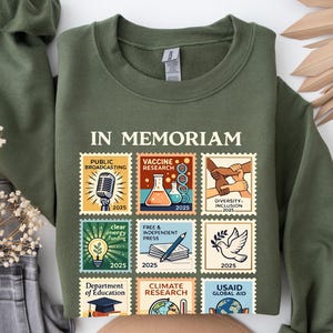 May include: Olive green sweatshirt with the words "IN MEMORIAM" above nine postage stamp-style graphics. The graphics depict themes like public broadcasting, vaccine research, diversity, and climate research. The year 2025 is printed on each stamp.
