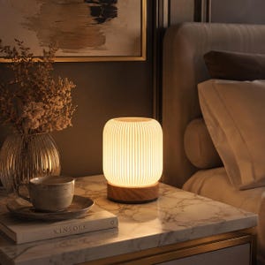 Op de afbeelding: Een cilindrische tafellamp met een witte, verticaal geribbelde kap en een ronde houten voet. De lamp verspreidt een warme, zachte gloed die de omgeving verlicht. Het ontwerp is eenvoudig en heeft een getextureerd oppervlak op de kap.