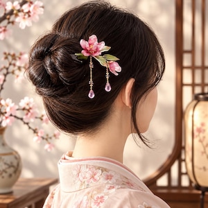 May include: A hair accessory featuring a pink flower with green leaves and two dangling gold chains, each ending in a small pink teardrop-shaped bead. The accessory is secured in a dark brown hair bun, showcasing detailed floral petals and delicate metal work.