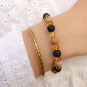 May include: A gold-colored bangle bracelet and a beaded bracelet with black, light brown, and amber-colored beads. The beaded bracelet features lava rock, wood, and amber beads. The bracelets are worn on a wrist, with a white eyelet shirt in the background.