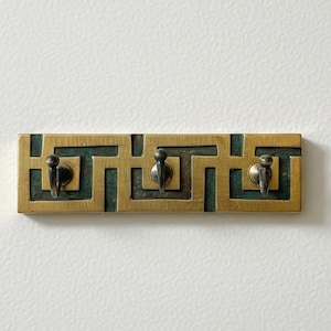 May include: A brass key holder with three hooks. The rectangular holder features a geometric design with a teal-colored background. The hooks are small and curved. The key holder is mounted on a white wall.