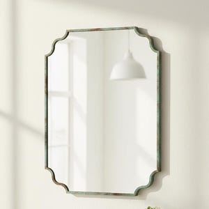 May include: A decorative wall mirror with a distressed green metal frame and a scalloped edge. The mirror hangs above a wooden shelf with books and a small potted plant. The room has a minimalist design.