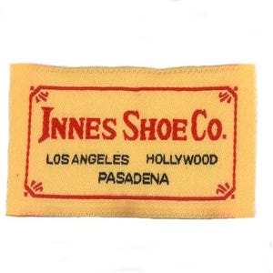 Op de afbeelding: Een geel geweven label met rode stiksels en rode tekst die "Innes Shoe Co. Los Angeles Hollywood Pasadena" luidt.