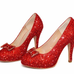 May include: A pair of red high heels covered in sparkling embellishments. Each shoe features a small bow with a square jewel. The heels are stiletto style and the inner sole is a light beige color.