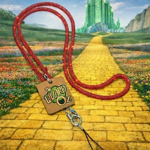 May include: A red glitter lanyard with a tag featuring "The Wizard of Oz" logo. The lanyard has a silver clasp and a black string. The background shows the yellow brick road leading to the Emerald City.
