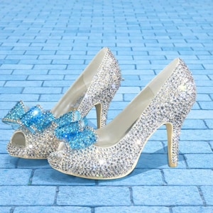 Puede incluir: Un par de tacones altos plateados cubiertos de brillantes pedrería. Cada zapato presenta una puntera abierta y un lazo azul claro. Los tacones se colocan sobre un fondo de ladrillos azules, creando un look glamuroso.