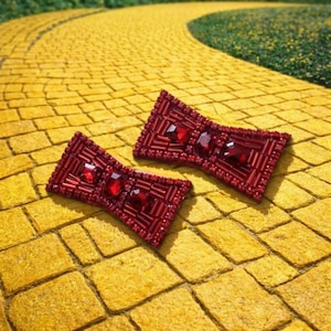 May include: Two red beaded bow-shaped hair clips with three red gems each. The clips are on a yellow brick road, with green grass in the background. The clips are likely for a costume or fashion accessory.