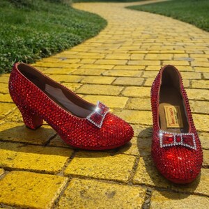 May include: A pair of sparkling red shoes covered in rhinestones. Each shoe has a small heel and a decorative bow with a silver-colored frame. The shoes are displayed on a yellow brick road.