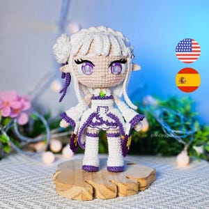 Pode incluir: Uma boneca de crochê feita à mão com cabelo branco, orelhas pontudas e grandes olhos roxos. A boneca veste uma roupa branca e roxa com um pequeno detalhe verde no pescoço. Ela está sobre uma base de madeira clara com textura orgânica.