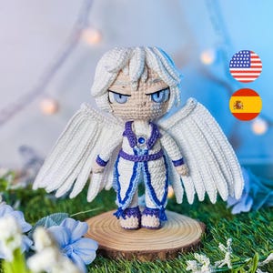 Pode incluir: Boneca de crochê feita à mão de um anjo com asas, cabelo e roupa brancos com detalhes em azul e roxo. A boneca tem olhos azuis e está em um disco de madeira. O fundo inclui flores azuis e vegetação.