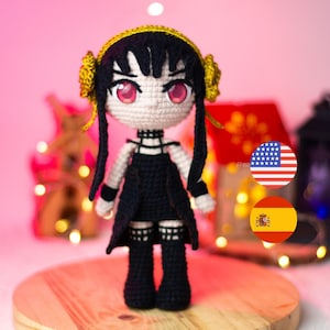 Pode incluir: Boneca de crochê feita à mão, representando uma personagem de Spy x Family. Possui cabelo preto, um vestido preto e detalhes dourados. A boneca tem olhos rosa e está sobre uma superfície de madeira com um fundo rosa. O design é detalhado.