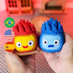 May include: Two handmade crochet figures, one red and yellow, the other blue, each with large white eyes and a smiling or neutral expression. The red figure has a flame-like top, while the blue figure has a spiky top. Both are held in hands.