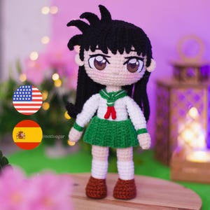 May include: A handmade crochet doll with black hair and large brown eyes. The doll wears a white shirt with a green collar and skirt, and brown shoes. The doll is standing on a wooden surface.