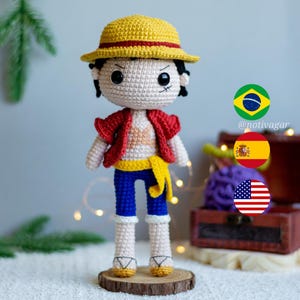 Puede incluir: Muñeca amigurumi de crochet con un sombrero de paja, una camisa roja, pantalones azules y un cinturón amarillo. La muñeca está de pie sobre una base de madera. La imagen también incluye las banderas de Brasil, España y los Estados Unidos.