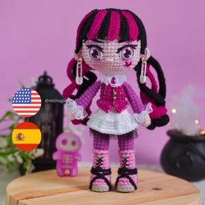 Op de afbeelding: Een handgemaakte gehaakte pop met een gotische uitstraling. De pop heeft zwart en roze haar, een paarse en roze outfit en roze laarzen. De pop heeft een witte rok en is versierd met veiligheidsspeld oorbellen.