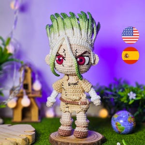 Pode incluir: Boneca de crochê feita à mão de um personagem com cabelo espetado verde e branco, olhos vermelhos e uma roupa bege. A boneca está em uma base de madeira. A boneca tem um cinto e uma pequena bolsa.