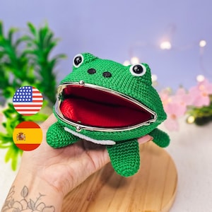 May include: A green crocheted frog-shaped coin purse with a red interior and silver-colored metal clasp. The frog has large, black and white eyes and small white feet. The purse is held open, revealing the red interior.