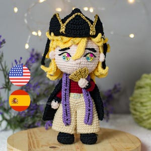 May include: Crocheted doll featuring a character with blonde curly hair, wearing a black hat with gold trim, a red shirt, and tan shorts. The doll has a purple scarf and is holding a gold fan. The doll is handmade.