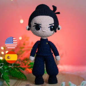 May include: A handmade crochet doll with black hair styled in a bun and dark blue outfit. The doll has large black eyes and is wearing a dark blue uniform. The doll is a unique, handcrafted item.
