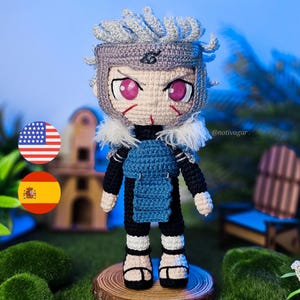 Puede incluir: Muñeca de ganchillo hecha a mano de un personaje con cabello gris, una diadema y un atuendo azul y negro. La muñeca tiene ojos rosados y está de pie sobre una rodaja de madera. El fondo es una escena borrosa al aire libre con vegetación.