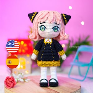 May include: A handmade crochet doll with pink hair, green eyes, and a black dress with gold trim. The doll is standing on a wooden surface. The doll has black shoes and white socks. The doll is a character from the anime series Spy x Family.