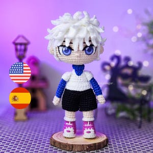 Pode incluir: Boneca de crochê feita à mão, com cabelo branco e olhos azuis. Usa uma camisa branca com mangas azuis, calções pretos e sapatos rosa e brancos. Está sobre uma base de madeira.