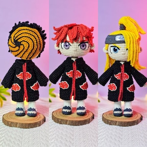 May include: Three handmade crochet dolls, each standing on a wooden base. The dolls feature black coats with red cloud designs, and distinct hairstyles: one with a spiral mask, one with red hair, and one with blonde hair.