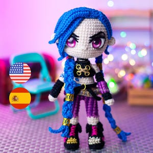 Puede incluir: Muñeca de ganchillo de un personaje con cabello azul en trenzas, con un top negro, pantalones morados y zapatos negros con detalles dorados. La muñeca tiene grandes ojos rosas y está sobre un fondo borroso. La muñeca mide aproximadamente 15 cm de altura.