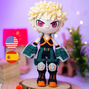 Puede incluir: Muñeco de ganchillo de un personaje con pelo rubio puntiagudo, ojos rojos y expresión decidida. El muñeco viste un atuendo negro y verde con detalles en naranja. El muñeco mide aproximadamente 20 cm de altura.