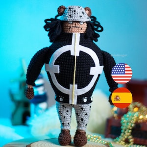 Pode incluir: Boneca de crochê feita à mão com um personagem em uma jaqueta preta com um design de alvo branco, calças cinza e brancas estampadas e um chapéu cinza com orelhas de urso marrons. A boneca tem cabelo castanho e sapatos marrons.