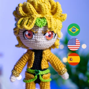 Peut inclure: Poupée amigurumi en crochet avec des cheveux jaunes, des yeux rouges et une chemise verte. La poupée porte une ceinture verte et a un cœur vert sur la poitrine. La poupée est debout devant un fond de drapeaux du Brésil, des États-Unis et de l'Espagne.