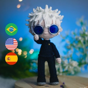 Op de afbeelding: Een gehaakte pop met wit haar, die een blauwe ronde bril en een zwarte outfit draagt. De pop staat op een houten oppervlak met een blauwe achtergrond.