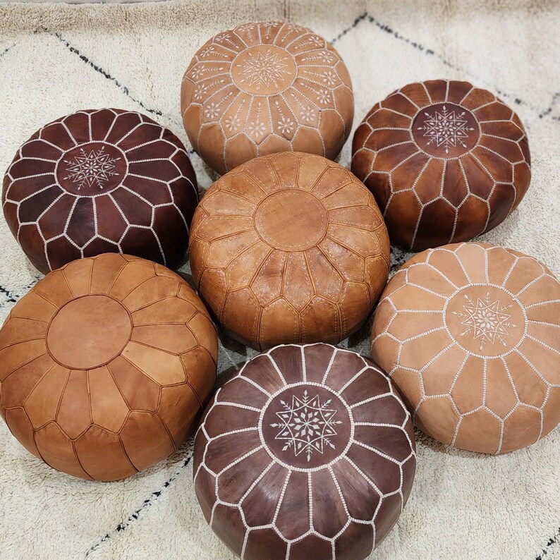 May include: Seven round leather poufs in various shades of brown and tan are arranged on a cream-colored rug. The poufs feature decorative stitching and patterns, with some having starburst designs. The leather appears to be soft and supple, suggesting a comfortable seating option.