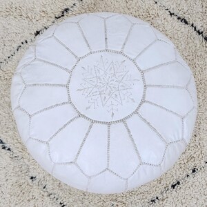 May include: A white leather Moroccan pouf with a snowflake design in the center. The round pouf is stitched with silver thread, and the snowflake is also embroidered with silver thread. The pouf is a decorative home furnishing.