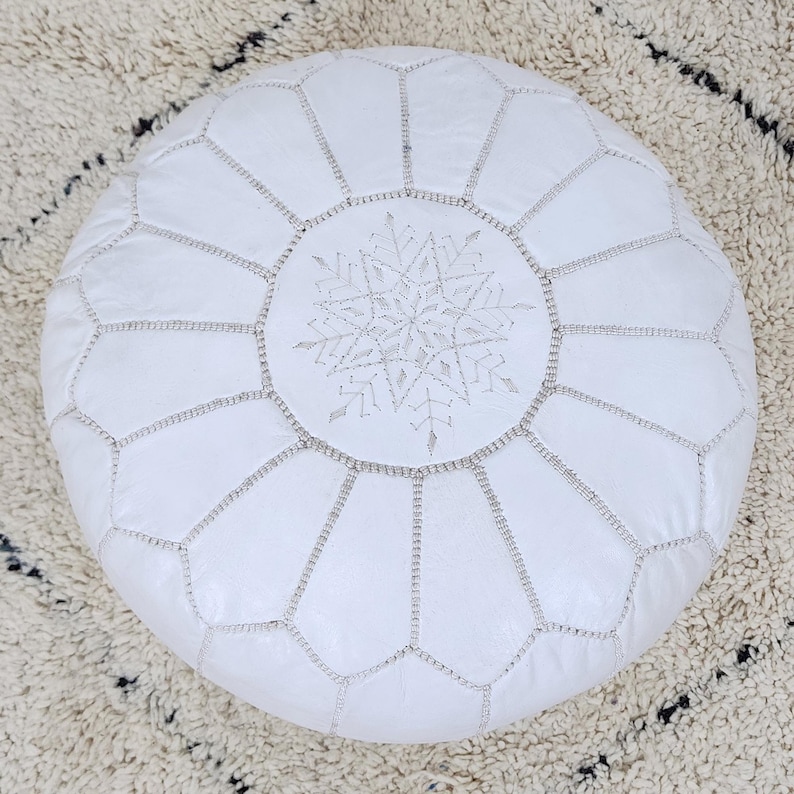 May include: A white leather Moroccan pouf with a snowflake design in the center. The round pouf is stitched with silver thread, and the snowflake is also embroidered with silver thread. The pouf is a decorative home furnishing.