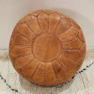 May include: Round, brown leather ottoman with white stitching. The ottoman has a circular design with a central circle and petal-like shapes radiating outwards. The stitching is visible and adds detail to the design. The ottoman is placed on a white rug.