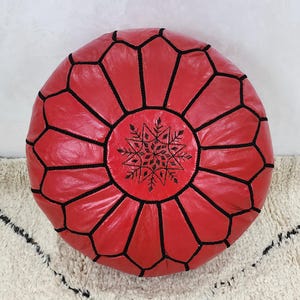 May include: A round, red leather pouf with black stitching and a central snowflake design. The pouf is a decorative floor cushion, perfect for adding a pop of color and texture to any living space. The design is geometric and the color is a vibrant red.