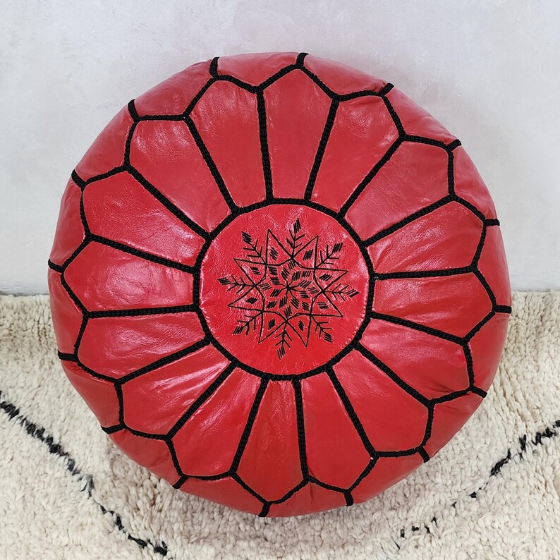 May include: A round, red leather pouf with black stitching and a central snowflake design. The pouf is a decorative floor cushion, perfect for adding a pop of color and texture to any living space. The design is geometric and the color is a vibrant red.