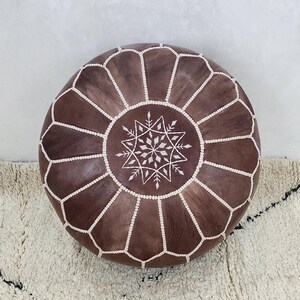 May include: A round, brown leather pouf with white stitching and a central starburst design. The pouf is a decorative piece of furniture, likely used as a footrest or extra seating. The design is geometric and the color is a rich brown.
