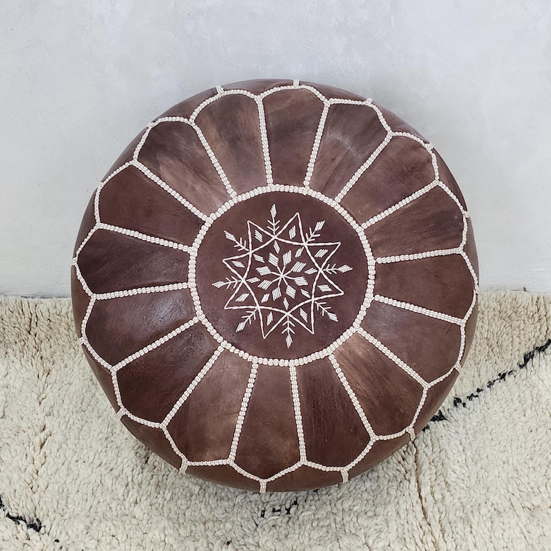 May include: A round, brown leather pouf with white stitching and a central starburst design. The pouf is a decorative piece of furniture, likely used as a footrest or extra seating. The design is geometric and the color is a rich brown.
