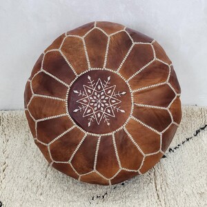 May include: Round brown leather ottoman with a geometric pattern and white stitching. The center features a white embroidered starburst design. The ottoman is placed on a textured white rug.