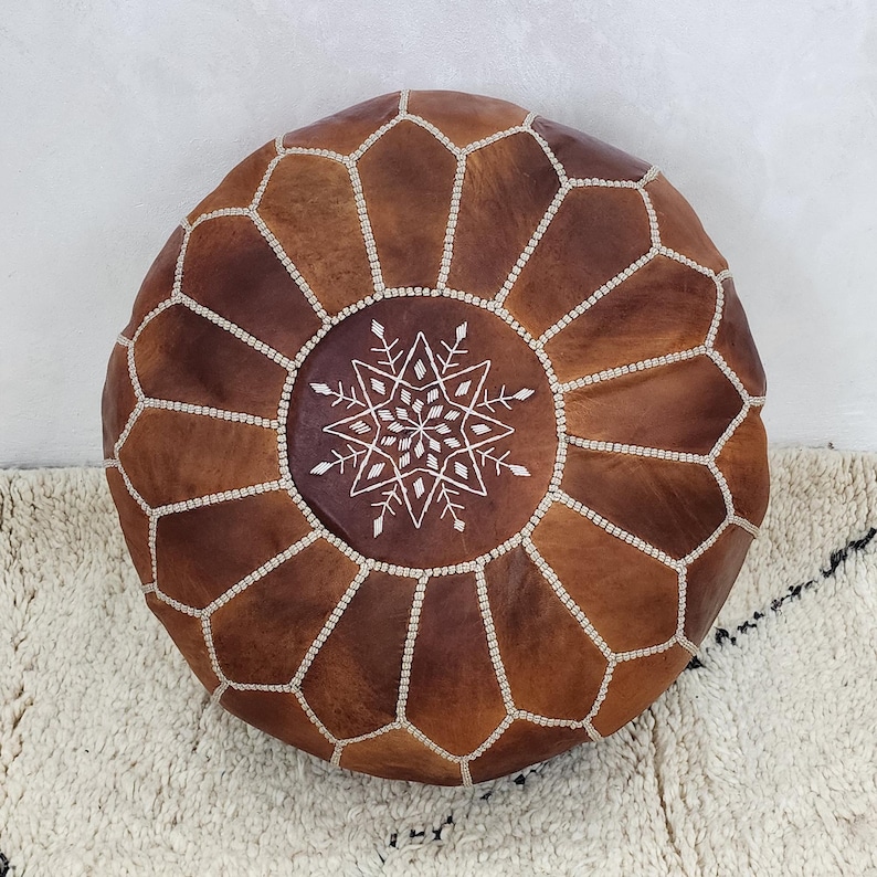 May include: Round brown leather ottoman with a geometric pattern and white stitching. The center features a white embroidered starburst design. The ottoman is placed on a textured white rug.